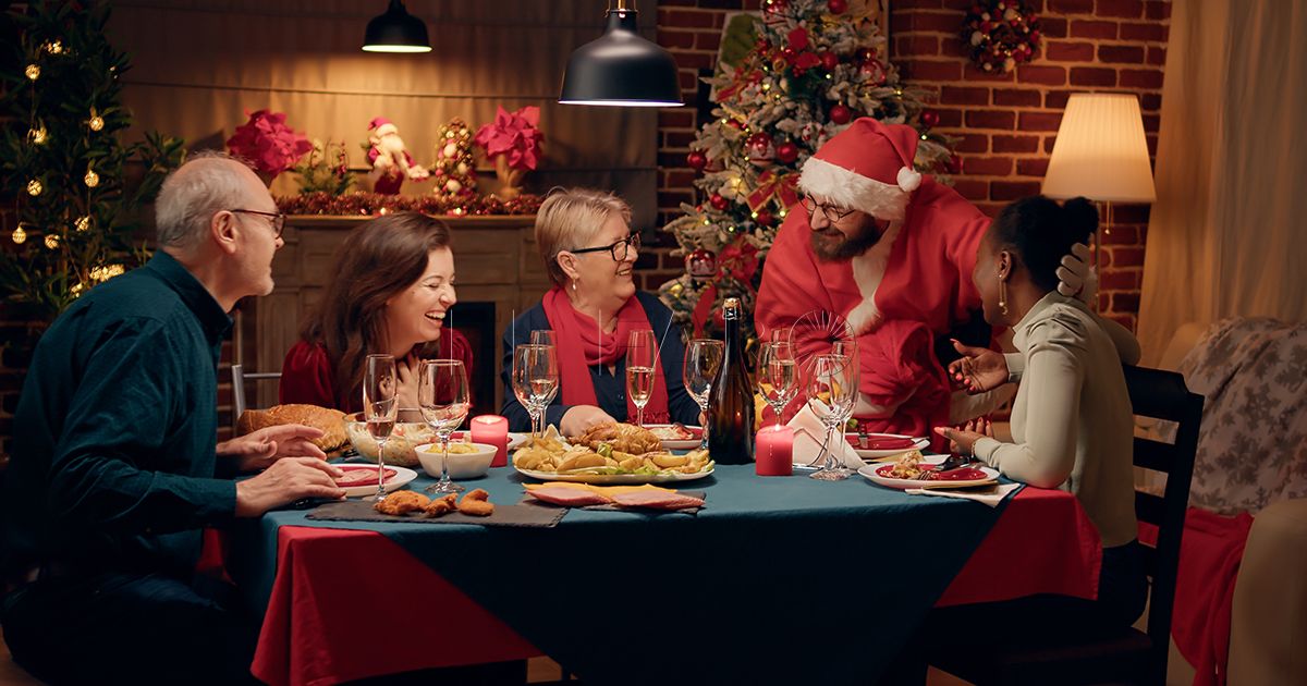 Man disguised as Santa Claus talking to happy woman at Christmas dinner at home. Cheerful african american woman surprised by husband looking like Santa while celebrating winter holiday.